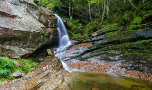 A serene waterfall cascades over rocky terrain, surrounded by lush greenery and a tranquil pool below.