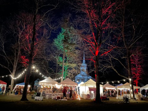 A festive night market with illuminated tents, trees, and a church steeple in the background, surrounded by bare branches.