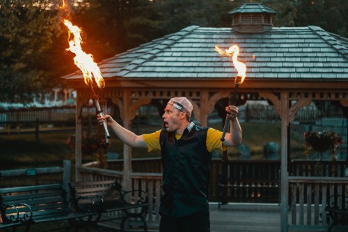 A performer in a yellow shirt and cap twirls flaming torches in a gazebo, showcasing a fire dance.