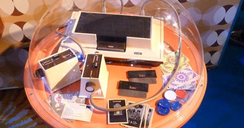 A vintage video game console and accessories displayed under a dome on an orange table, surrounded by retro patterns.
