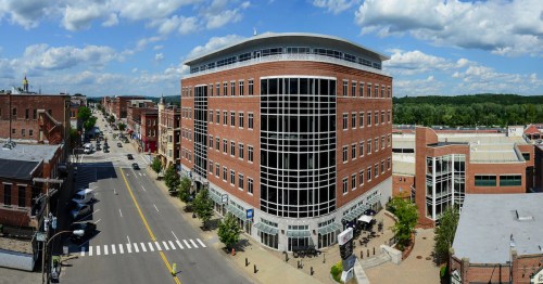 A panoramic view of a brick building in a town, with trees lining the street and a blue sky filled with clouds.