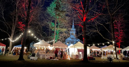 A festive night market with illuminated tents, trees lit in red and green, and a church steeple in the background.