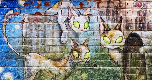 Colorful mural of three cats with bright green eyes, set against a textured brick wall.