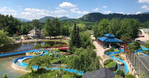 A scenic view of a water park with slides, a pond, and lush greenery, set against a backdrop of mountains.