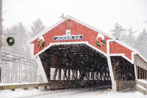 A red covered bridge in Jackson, NH, adorned with wreaths, surrounded by falling snow in a winter landscape.