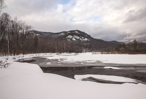 A snowy landscape with a river, surrounded by trees and mountains under a cloudy sky.