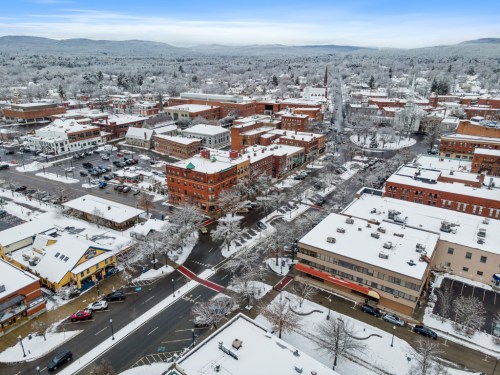 Aerial view of a snowy town with buildings, streets, and trees blanketed in white, under a clear blue sky.