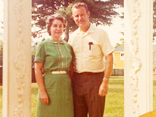 A couple stands together in a park, framed by an ornate white structure, smiling in vintage clothing.