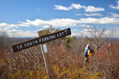 A wooden sign pointing to the south parking lot, with hikers in the background and a clear blue sky.