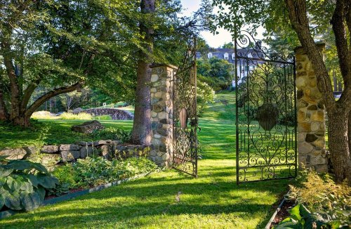 A wrought iron gate opens to a lush green garden with trees and stone walls, leading to a distant house.