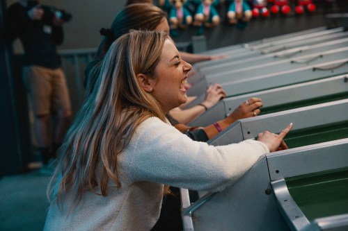 A woman with long hair excitedly plays a game, focused and cheering, with friends in the background.