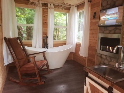 Cozy wooden interior featuring a rocking chair, freestanding bathtub, and a TV beside a stone fireplace.