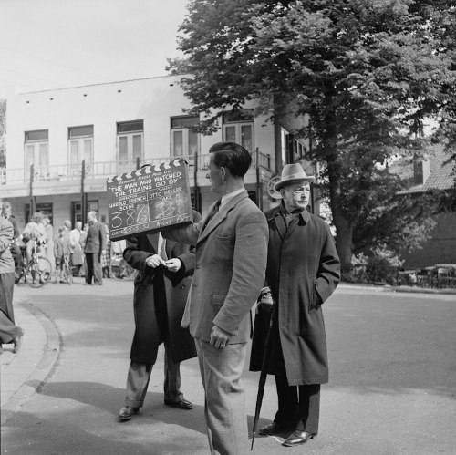 A group of men in vintage clothing, one holding a film slate, on a street with buildings in the background.