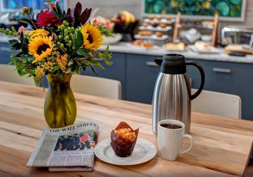A cozy breakfast scene with a vase of flowers, coffee, a muffin, a newspaper, and a thermos on a wooden table.