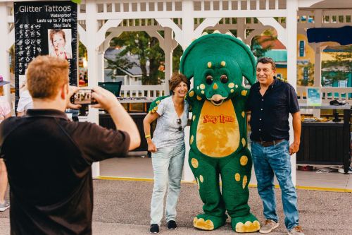 A person in a dinosaur costume poses with two adults for a photo at an outdoor event.
