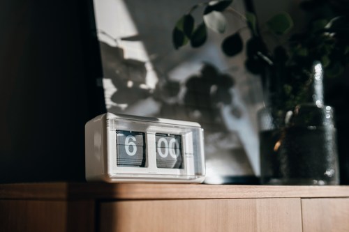 A retro flip clock displaying 6:00, resting on a wooden surface with shadows and a plant in the background.