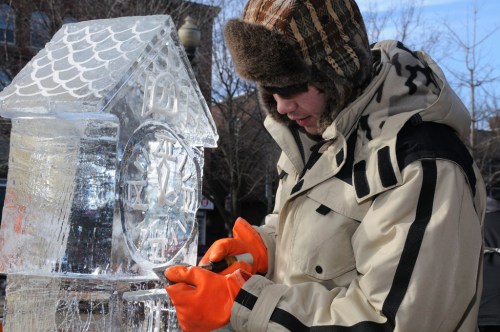 A person in a winter coat and hat carves an ice sculpture of a clock, wearing bright orange gloves.