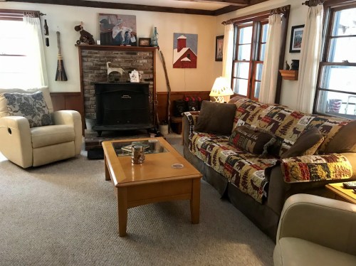 Cozy living room with a sofa, armchair, coffee table, and a fireplace, decorated with artwork and a quilt.