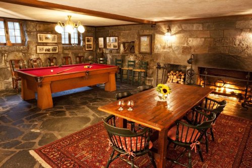 Cozy game room with a pool table, wooden dining table, chairs, and a fireplace, featuring stone walls and a warm atmosphere.