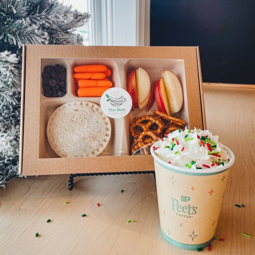 A snack box with carrots, apple slices, pretzels, and chocolate chips next to a festive coffee cup topped with whipped cream.