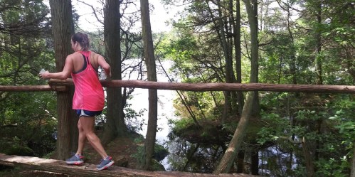 A person in a pink tank top walks along a log bridge in a forest, with a serene lake visible in the background.