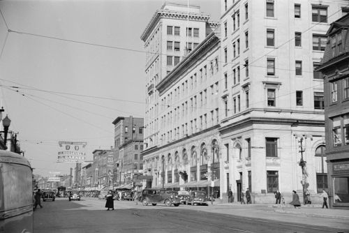 Historic black-and-white photo of a busy city street with vintage cars, buildings, and pedestrians.