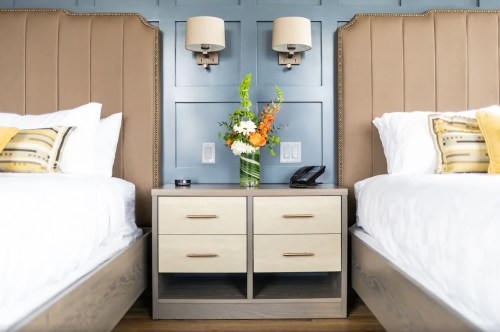 A stylish hotel room with two beds, a nightstand, and decorative flowers against a blue wall.