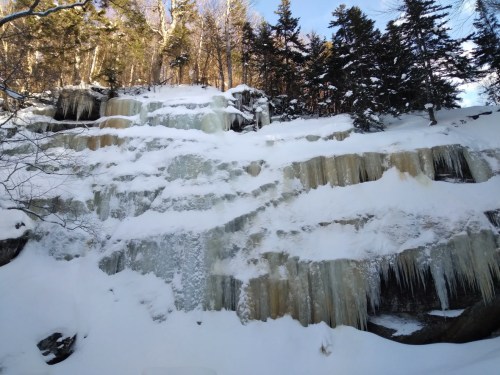 A snowy landscape featuring a frozen waterfall cascading down a rocky cliff, surrounded by trees.