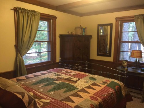 Cozy bedroom with a quilted bedspread, wooden furniture, and large windows overlooking a forested view.