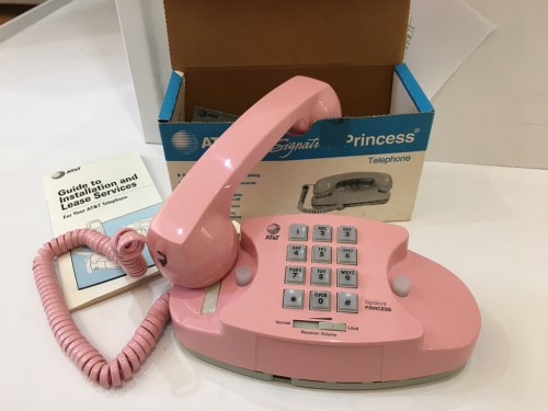A pink Princess telephone with a coiled cord, next to its box and an instruction manual.