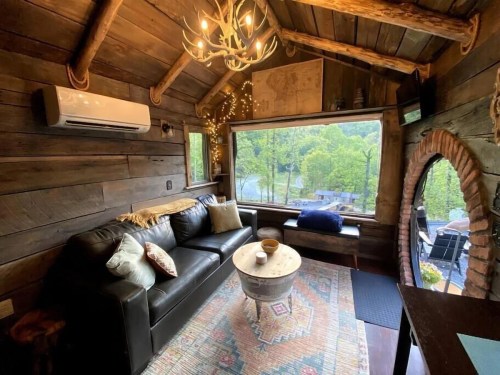 Cozy cabin interior with a leather sofa, rustic decor, and large windows overlooking a scenic view.
