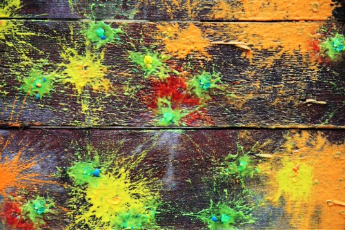 Colorful splatters of orange, yellow, and green paint on a textured wooden surface.