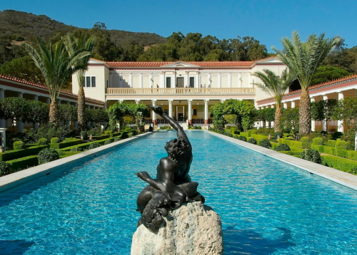 A serene pool with a statue in the foreground, surrounded by lush greenery and a grand building in the background.