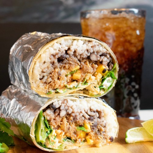 A close-up of a cut burrito filled with rice, beans, meat, and vegetables, alongside a drink and a lemon wedge.