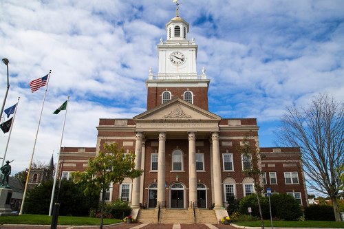 A brick municipal building with a clock tower, flanked by flags and surrounded by trees under a cloudy sky.