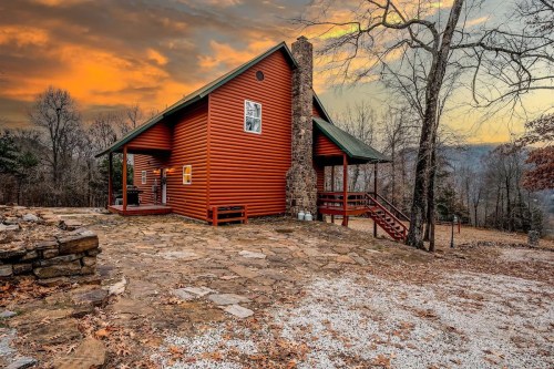 A cozy wooden cabin with a stone chimney, surrounded by trees and a rocky path, under a colorful sunset sky.