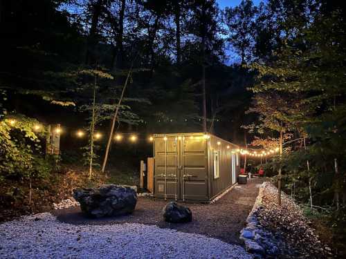 A cozy container home illuminated by string lights, surrounded by trees and gravel pathways at dusk.