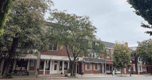 A quiet street lined with brick buildings and trees under a cloudy sky. Shops and cafes are visible along the sidewalk.