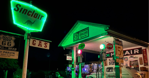 A vintage gas station at night, illuminated by neon signs, featuring a Sinclair dinosaur logo and retro decor.