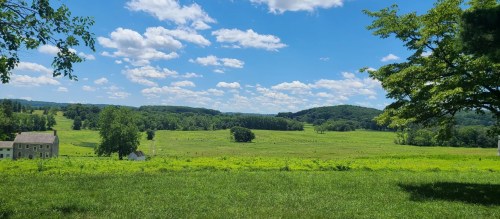 A scenic landscape featuring green fields, rolling hills, and a bright blue sky with fluffy white clouds.