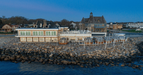 A coastal building with a deck and glass structures, surrounded by rocks and water, under a clear blue sky.