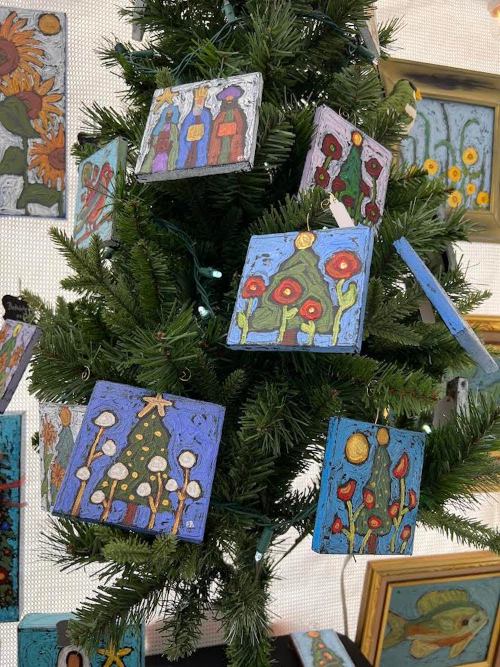 Colorful handmade ornaments hang from a green Christmas tree, featuring various artistic designs and patterns.