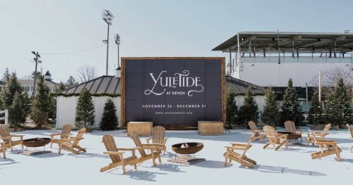 A winter scene featuring a large sign for "Yuletide at Devon" surrounded by wooden chairs and evergreen trees.
