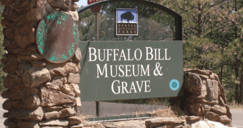 Sign for the Buffalo Bill Museum & Grave, featuring stone pillars and a green background in a natural setting.