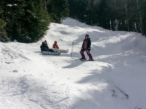 Three people enjoying a snowy slope, with one standing on a snowboard and two sitting on a sled. Trees in the background.