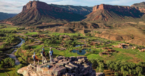 Three hikers stand on a rocky outcrop, overlooking a lush valley with mountains in the background and a winding river.
