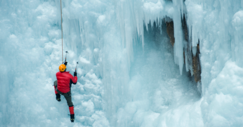 A climber in a red jacket and yellow helmet ascends a frozen ice wall with icicles hanging above.