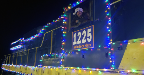 A decorated train engine with colorful lights, featuring a person in a winter hat smiling from the window.