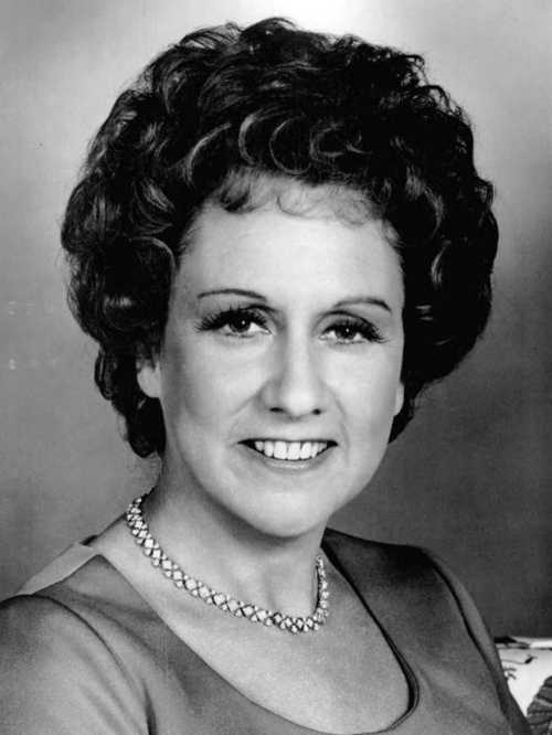 Black and white portrait of a smiling woman with curly hair, wearing a necklace and a dress, against a neutral background.