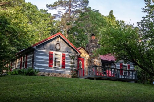 A charming log cabin with red shutters and a porch, surrounded by lush greenery and trees.